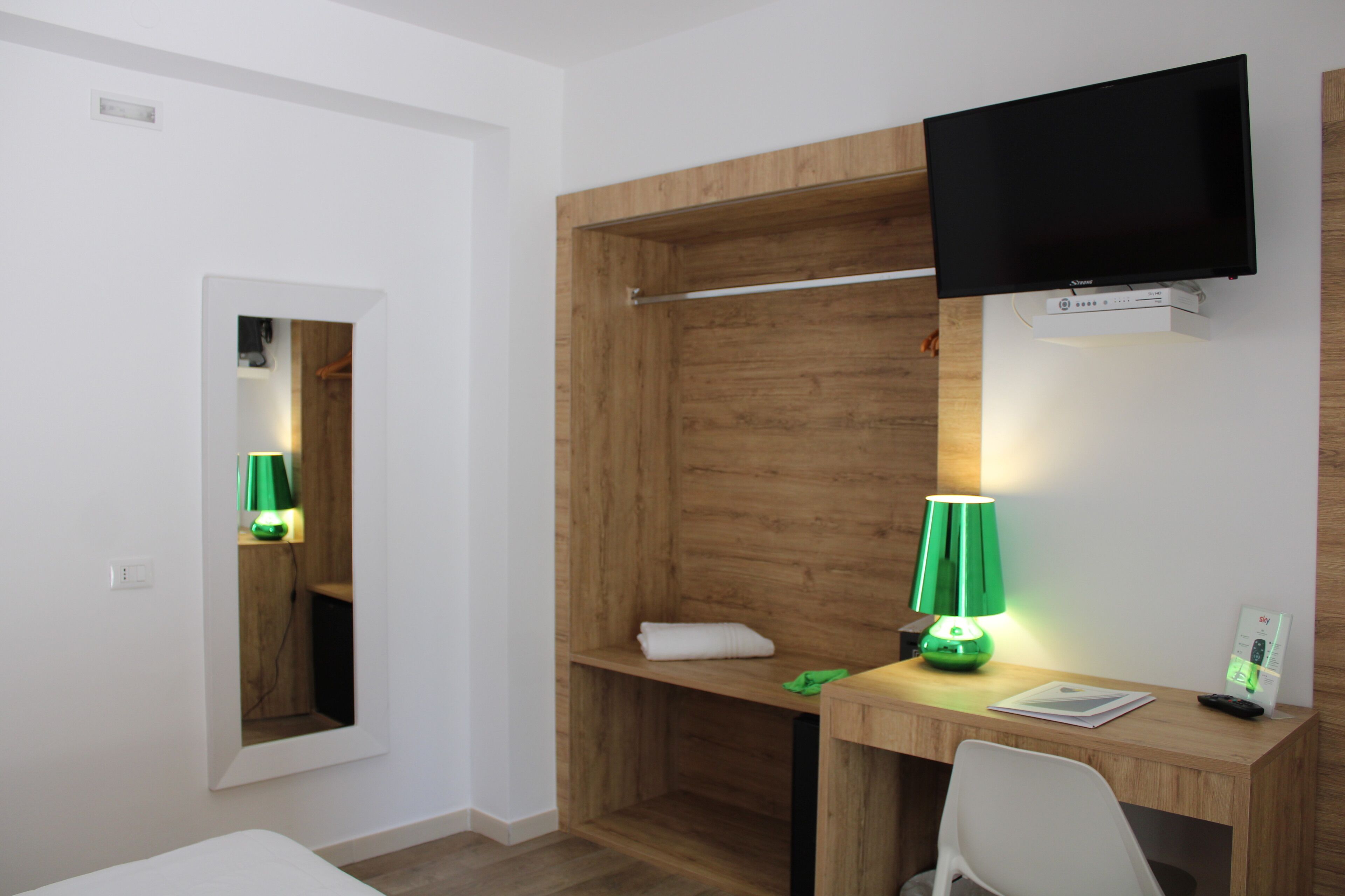 A hotel room with a wooden desk, a television mounted on the wall, a green desk lamp, a white chair, and a small shelf with a towel and a green cloth.