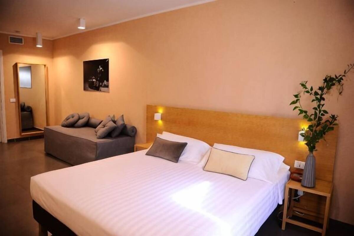 A hotel room with a large bed, a sofa, a nightstand with a vase, and a mirror.