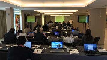 A conference room with attendees seated at tables with laptops, a presentation screen, and a whiteboard.