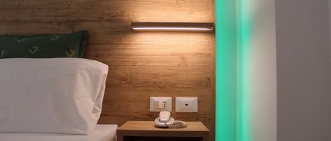 A neatly made bed with white linens, a wooden headboard, and a bedside table with a lamp and a power outlet.