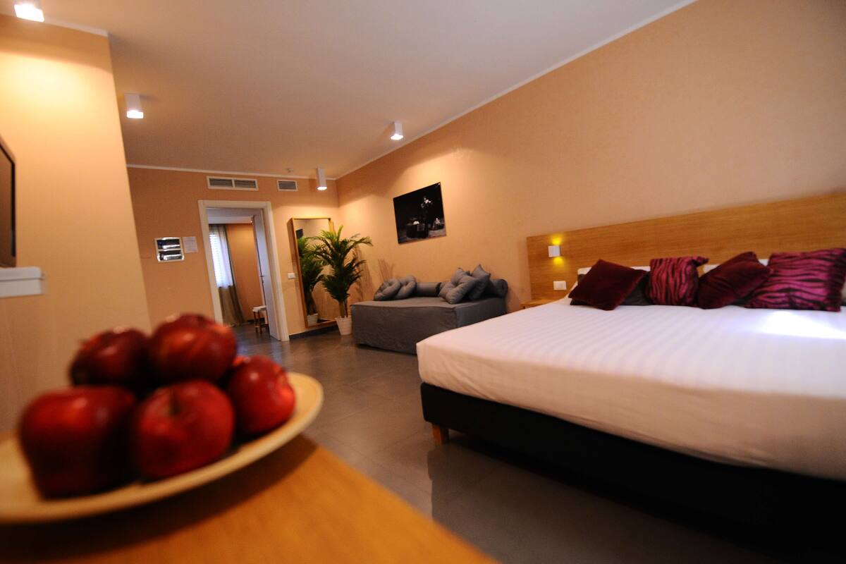 A hotel room with a bed, a sofa, a small table with apples, and a plant.