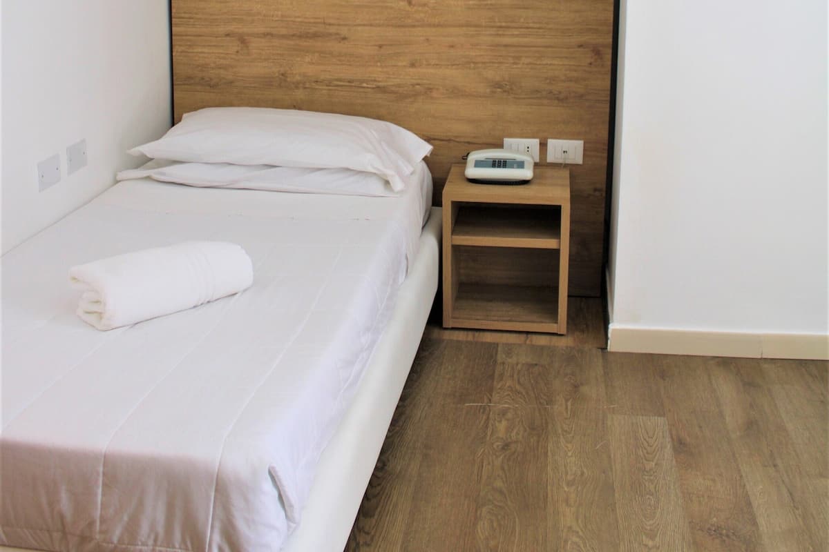 A small, neatly made bed with a wooden headboard in a minimalistic room with wooden flooring and white walls.