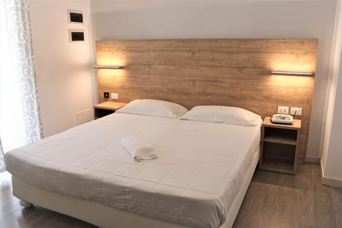 Una cama bien hecha con cabecera de madera, mesitas de noche con lámparas y una toalla blanca sobre la cama.