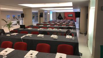 Un salón de conferencias con filas de mesas, sillas rojas y una zona de escenario.