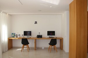 Un ufficio moderno con due scrivanie, ciascuna dotata di monitor, tastiera e mouse. Due sedie nere con gambe in legno. Un simbolo Wi-Fi è visibile sulla parete.