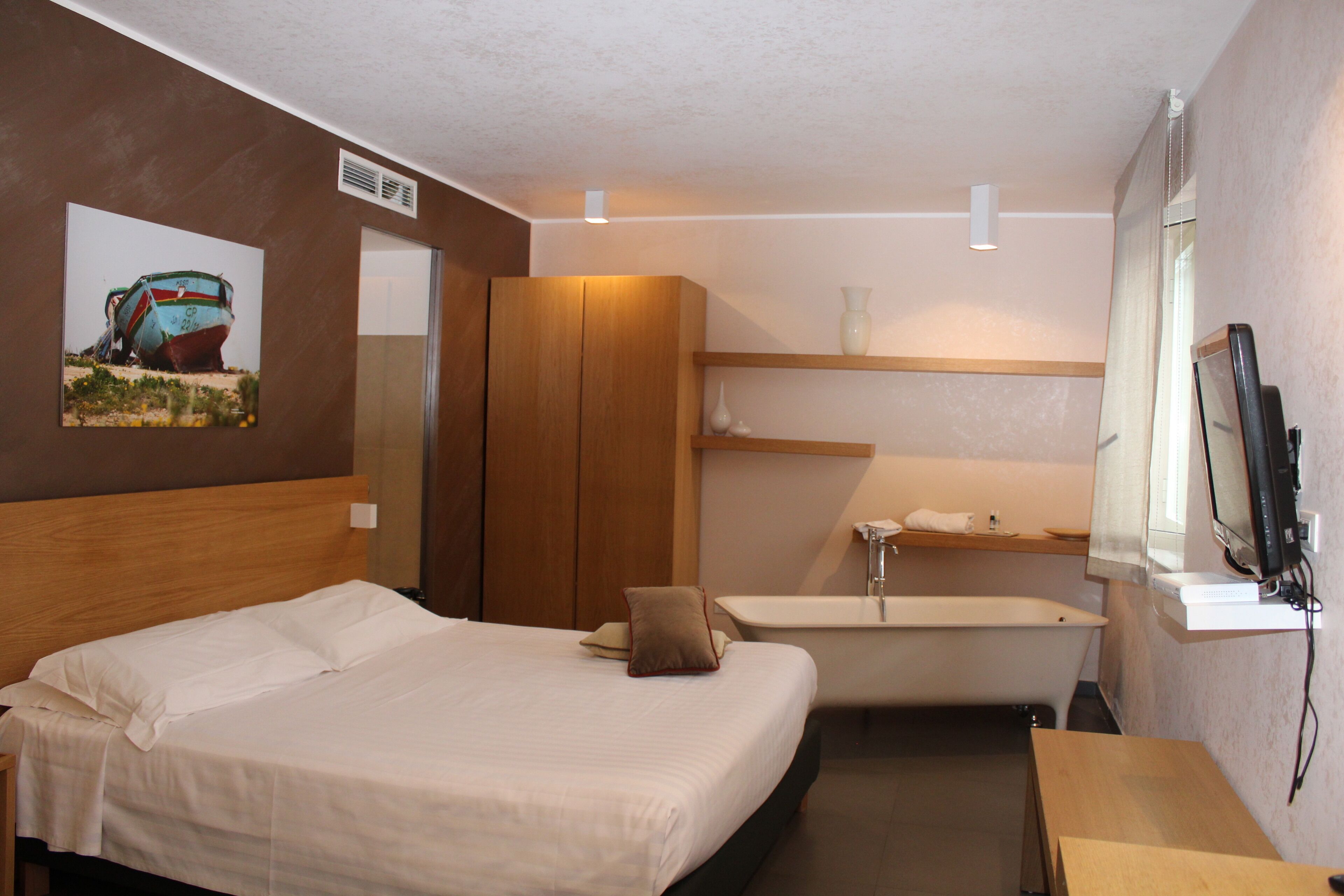 A hotel room with a bed, a wooden wardrobe, a bathroom with a bathtub, and a TV mounted on the wall.