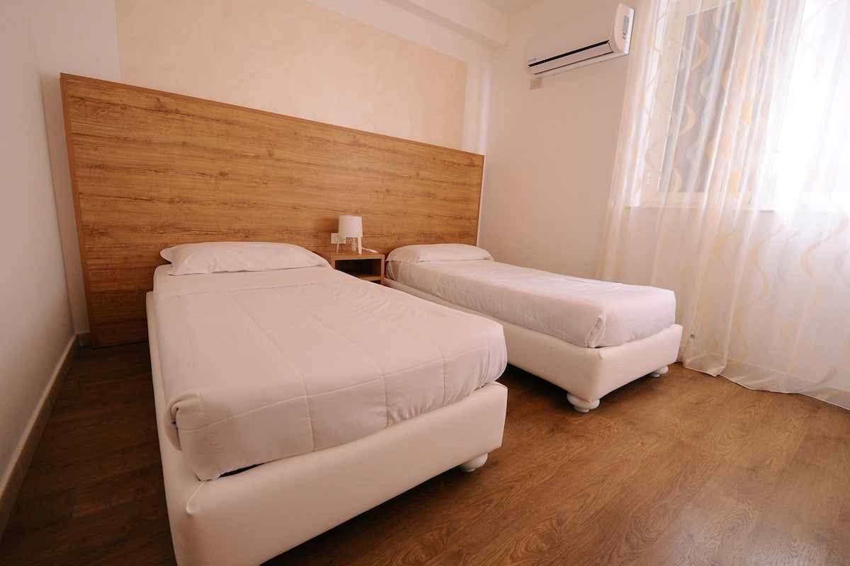 A room with two beds, a wooden headboard, a nightstand with a lamp, and a window with sheer curtains.