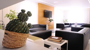 A modern living room with a large cactus in a woven basket, black sofas, a flat-screen TV mounted on a wooden wall, and a small table with books. - Casena Dei Colli, Sure Hotel Collection by Best Western (Palermo)