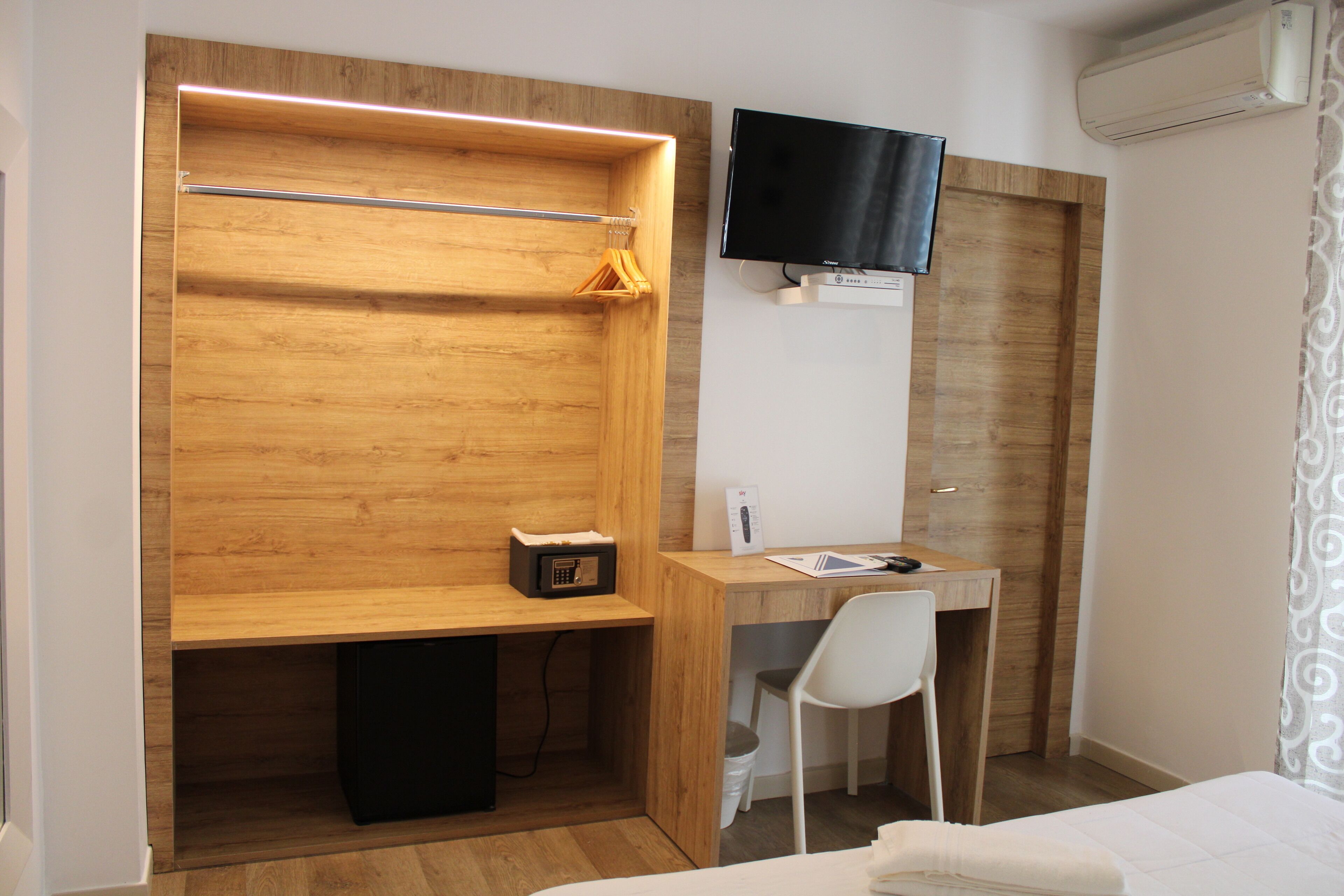 Habitación de hotel compacta con armario de madera, un escritorio con silla, cama, televisor montado en la pared y equipo de aire acondicionado.