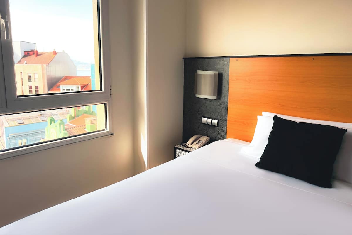 Une chambre d’hôtel équipée d’un lit, d’un téléphone sur la table de chevet, d’une lampe fixée au mur et d’une fenêtre donnant sur des immeubles.