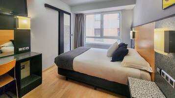 A modern hotel room with a bed, a desk, a chair, a window, and a bathroom.