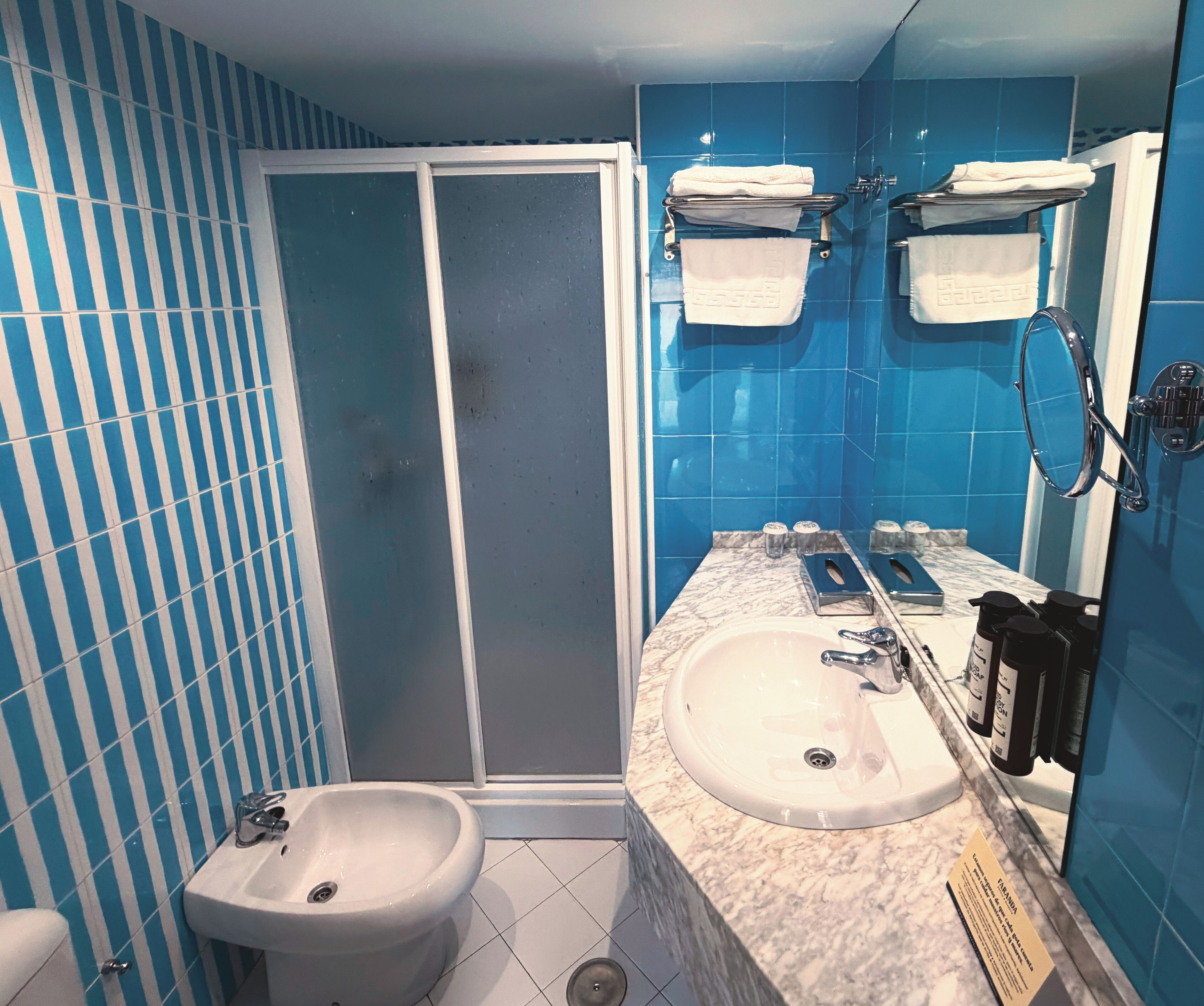 Un baño con un lavamanos blanco, un bidé, una ducha con puerta de vidrio y paredes de azulejos azules.