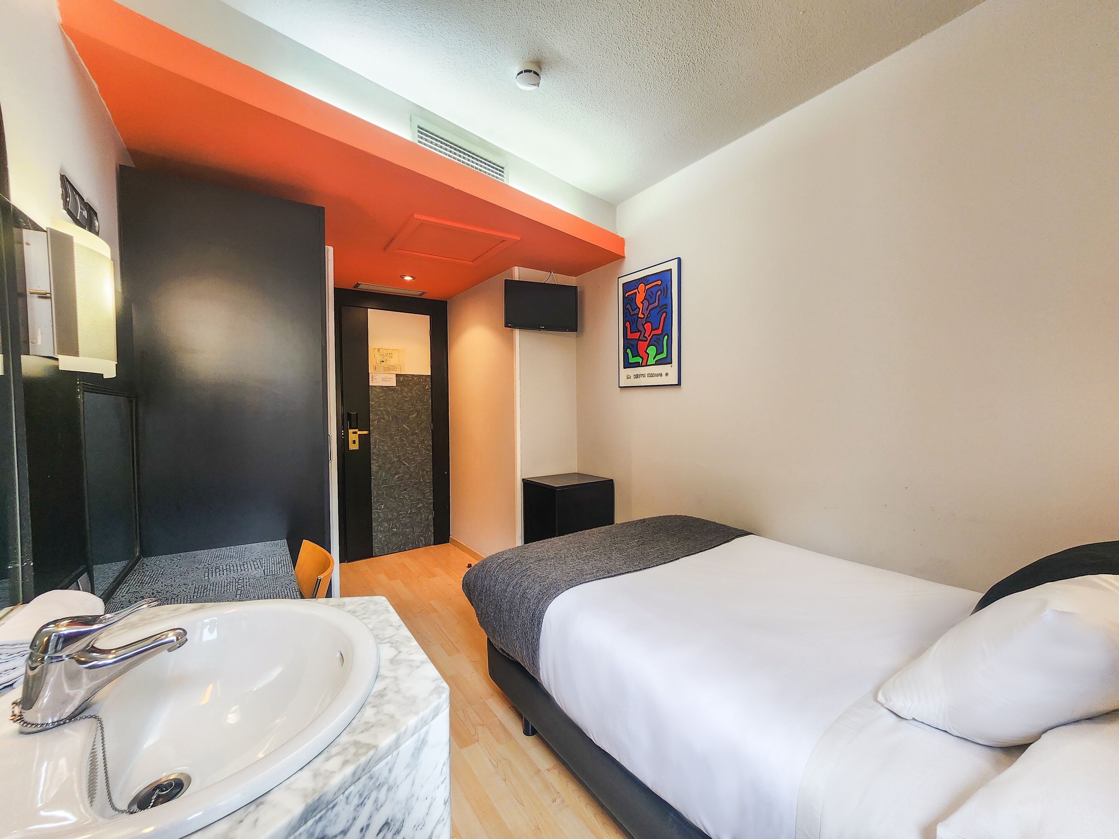 A modern hotel room with a marble sink, a bed with white bedding, a black headboard, a colorful abstract painting, and a bright orange accent wall.