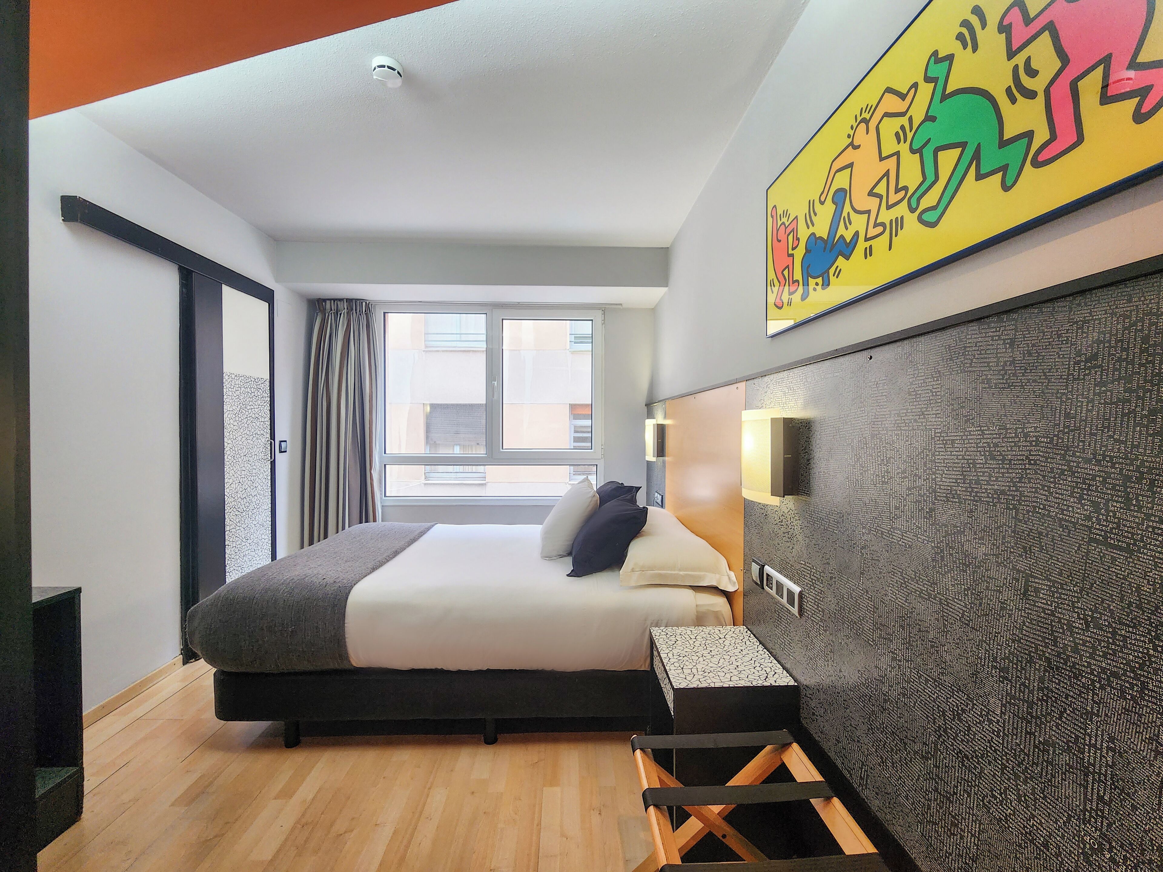 A modern hotel room with a large bed, a small side table, a decorative wall, and a window with curtains.