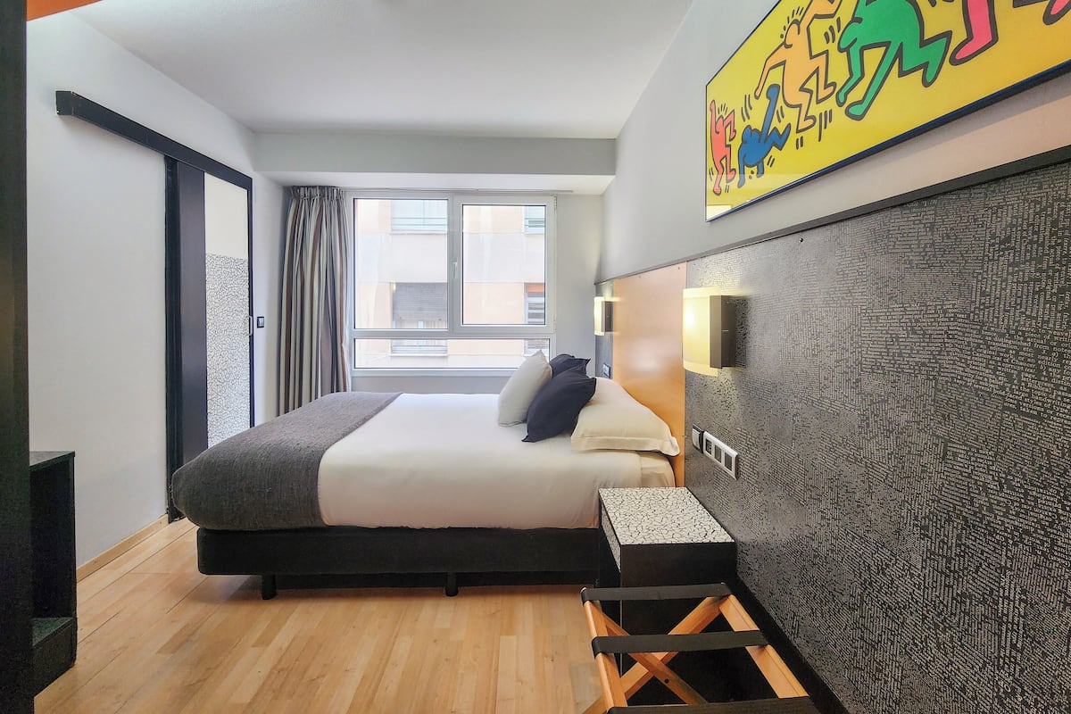 Une chambre d’hôtel moderne avec un grand lit, une petite table de chevet, un mur décoratif et une fenêtre avec des rideaux.