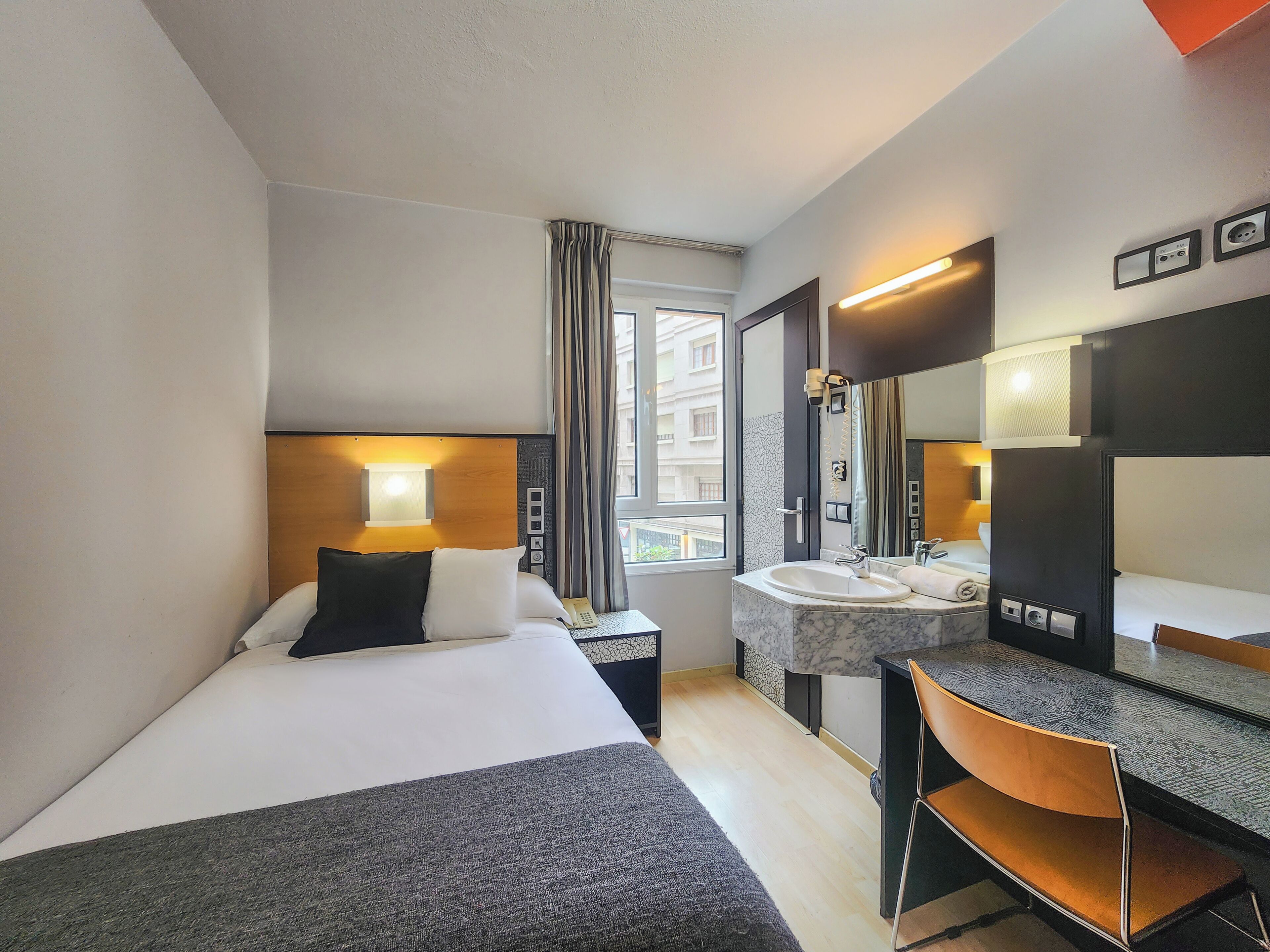 A hotel room with a bed, a desk, a chair, a bathroom with a sink and mirror, and a window with curtains.