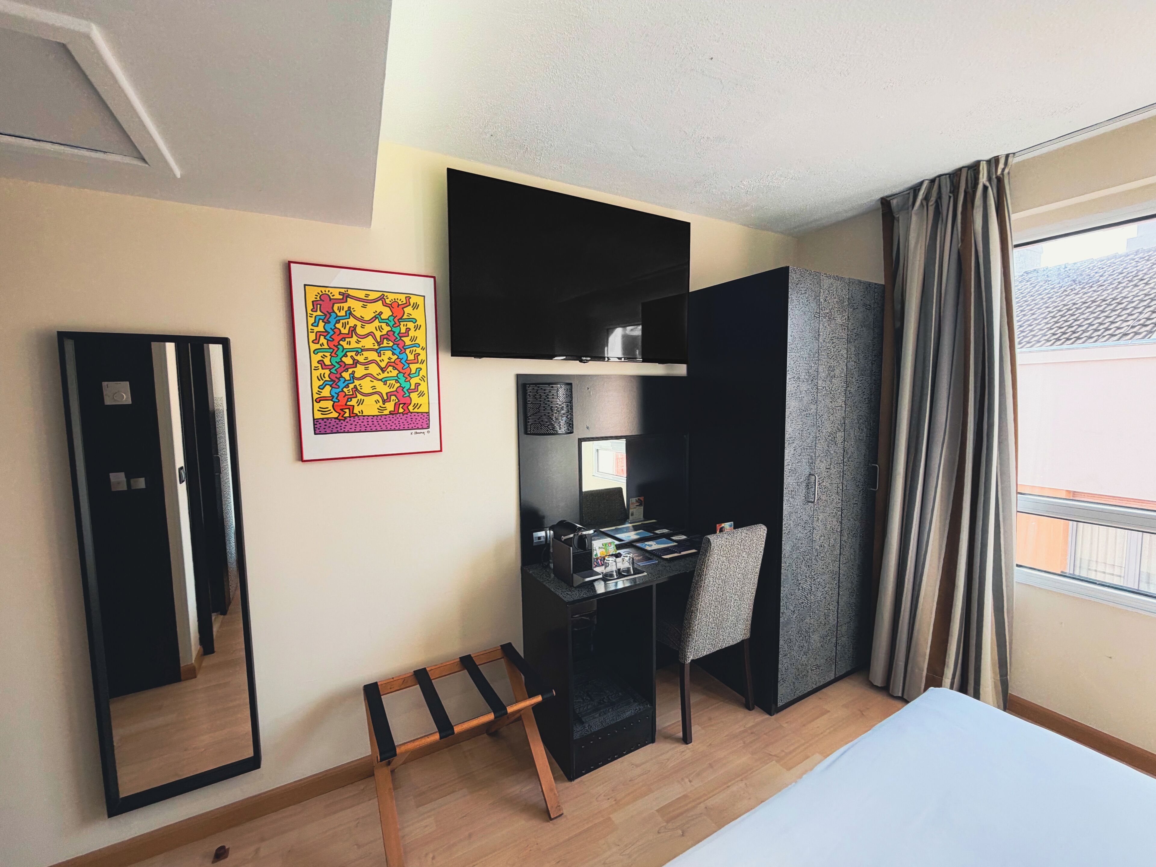 Hotelkamer met een bed, een bureau met stoel, een spiegel, een lijst met foto en een raam met gordijnen.