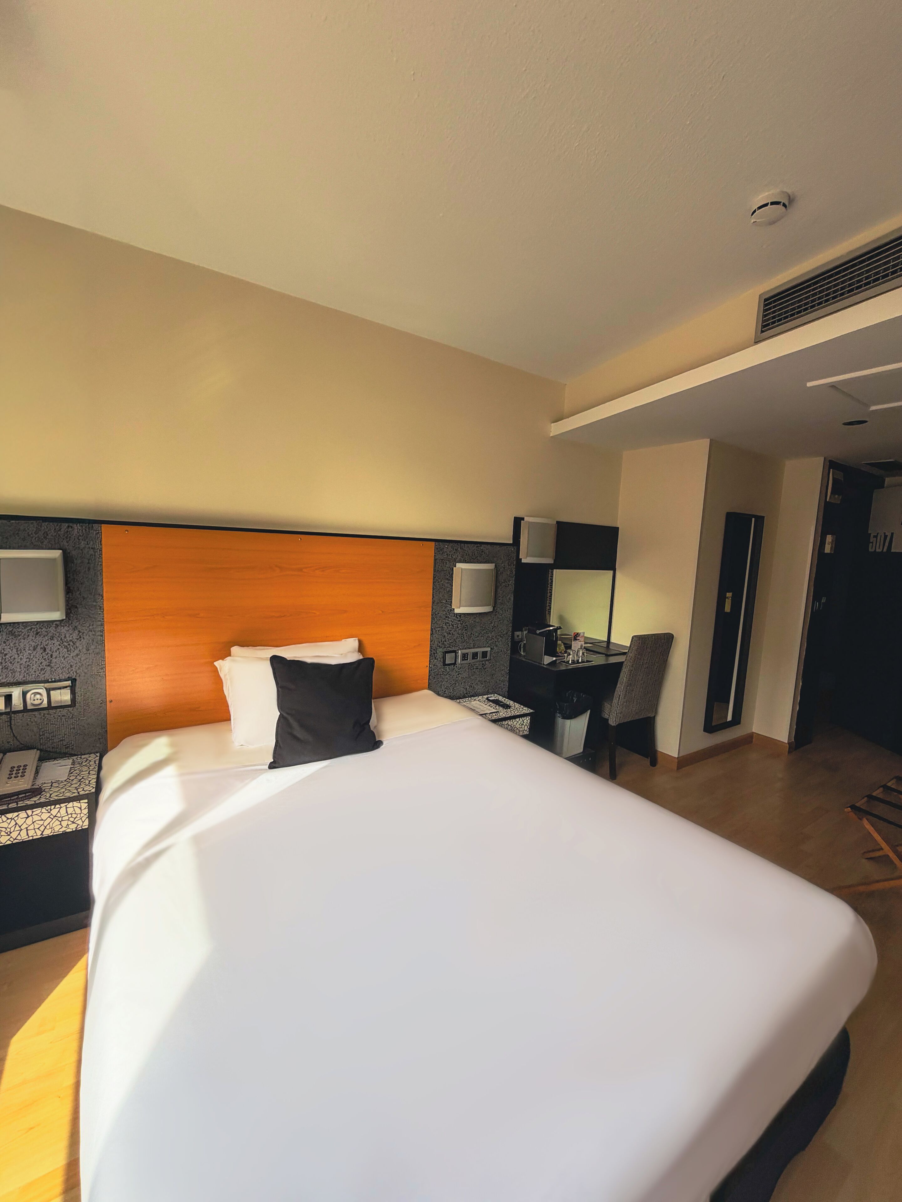 Quarto de hotel com uma cama grande, uma escrivaninha com cadeira, telefone, espelho e uma pequena mesa.