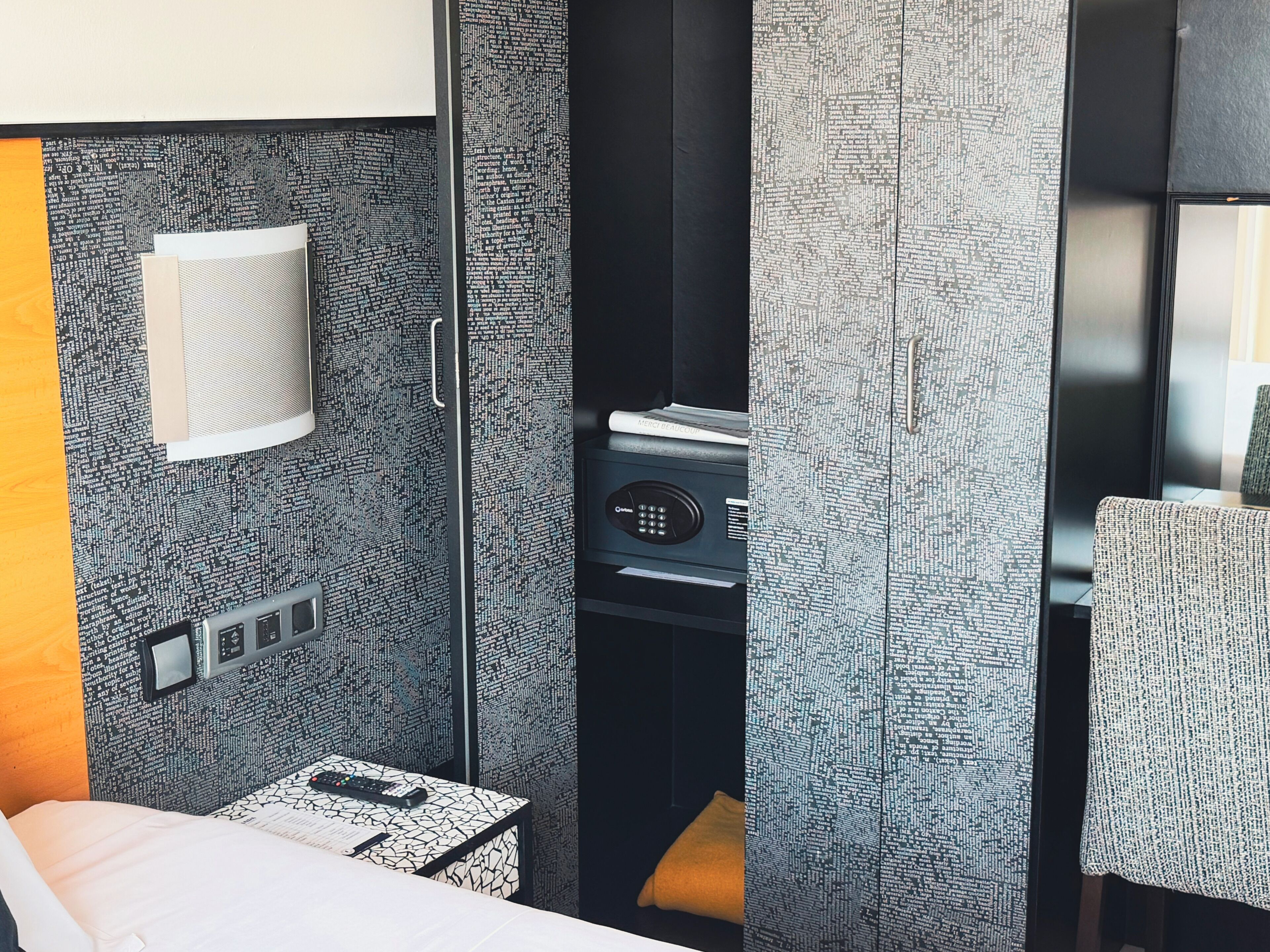A modern hotel room with a bed, a desk, a chair, a small table, a wall-mounted control panel, and a large window.