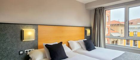 A hotel room with two beds, a wooden headboard, a nightstand, and a large window with a view of buildings.