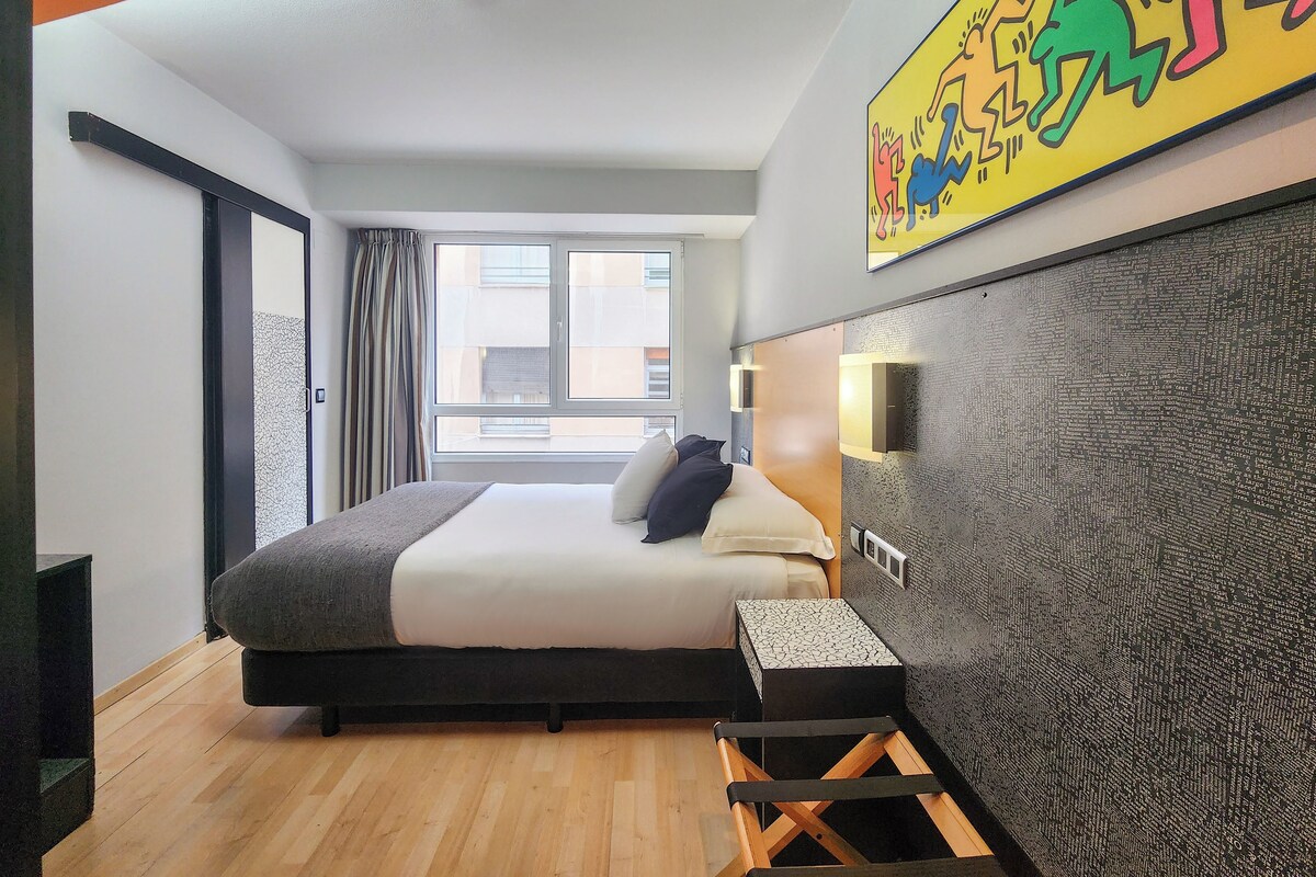 Ein modernes Hotelzimmer mit einem großen Bett, einem kleinen Beistelltisch, einer Wanddekoration und einem mit Vorhängen versehenen Fenster.