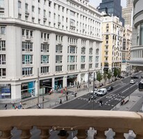 A multi-story building with large windows and balconies, a street with cars and pedestrians, and a pedestrian crossing.