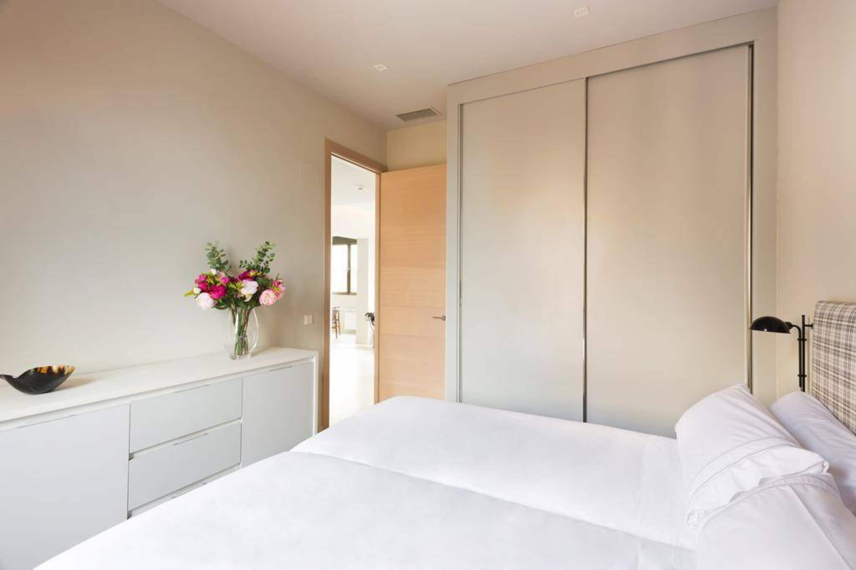 Un dormitorio moderno con una cama, una mesita de noche con un florero y un armario, con vista a otra habitación.
