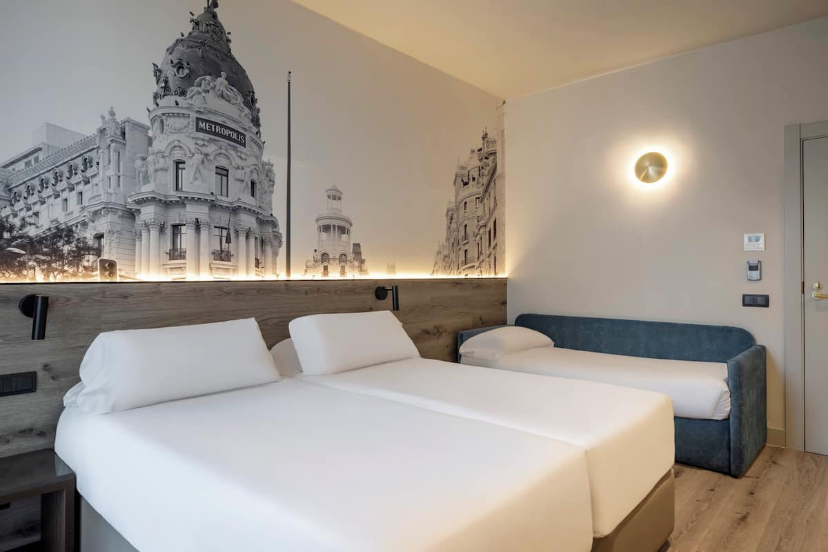 Une chambre d’hôtel avec deux lits, une grande fresque représentant un bâtiment, une tête de lit en bois et une table de chevet.