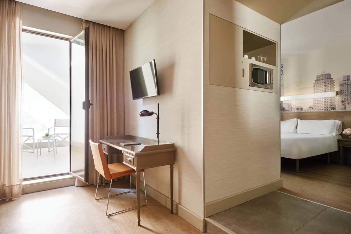 Une chambre d’hôtel équipée d’un bureau, d’une chaise, d’un lit, d’une télévision et offrant une vue sur le paysage urbain par la fenêtre.