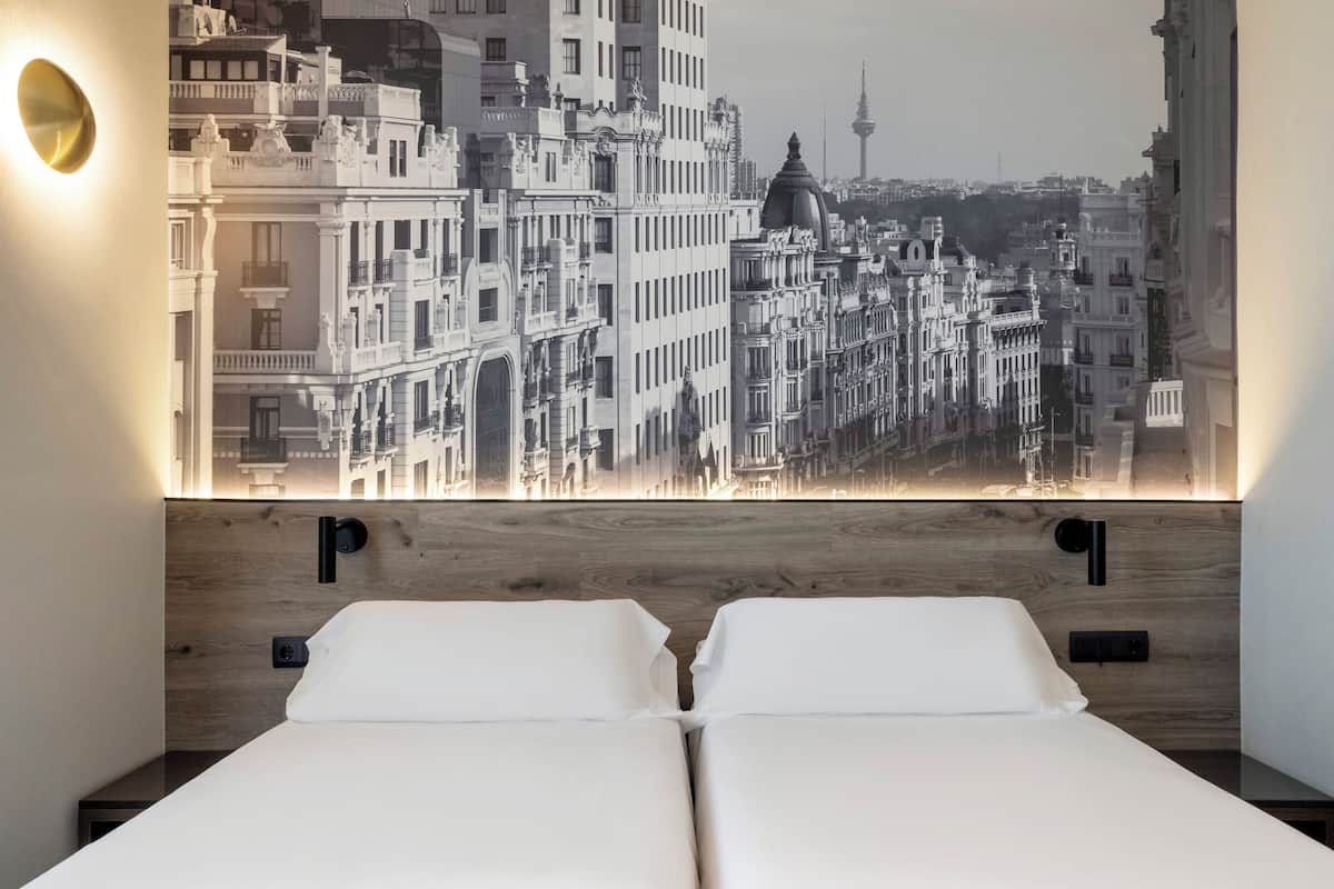 Une chambre d’hôtel avec une grande fresque représentant un paysage urbain sur le mur, deux lits et un luminaire moderne.