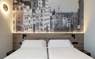 Ein Hotelzimmer mit großem Stadtbild-Wandgemälde, zwei Betten und einer modernen Leuchte.