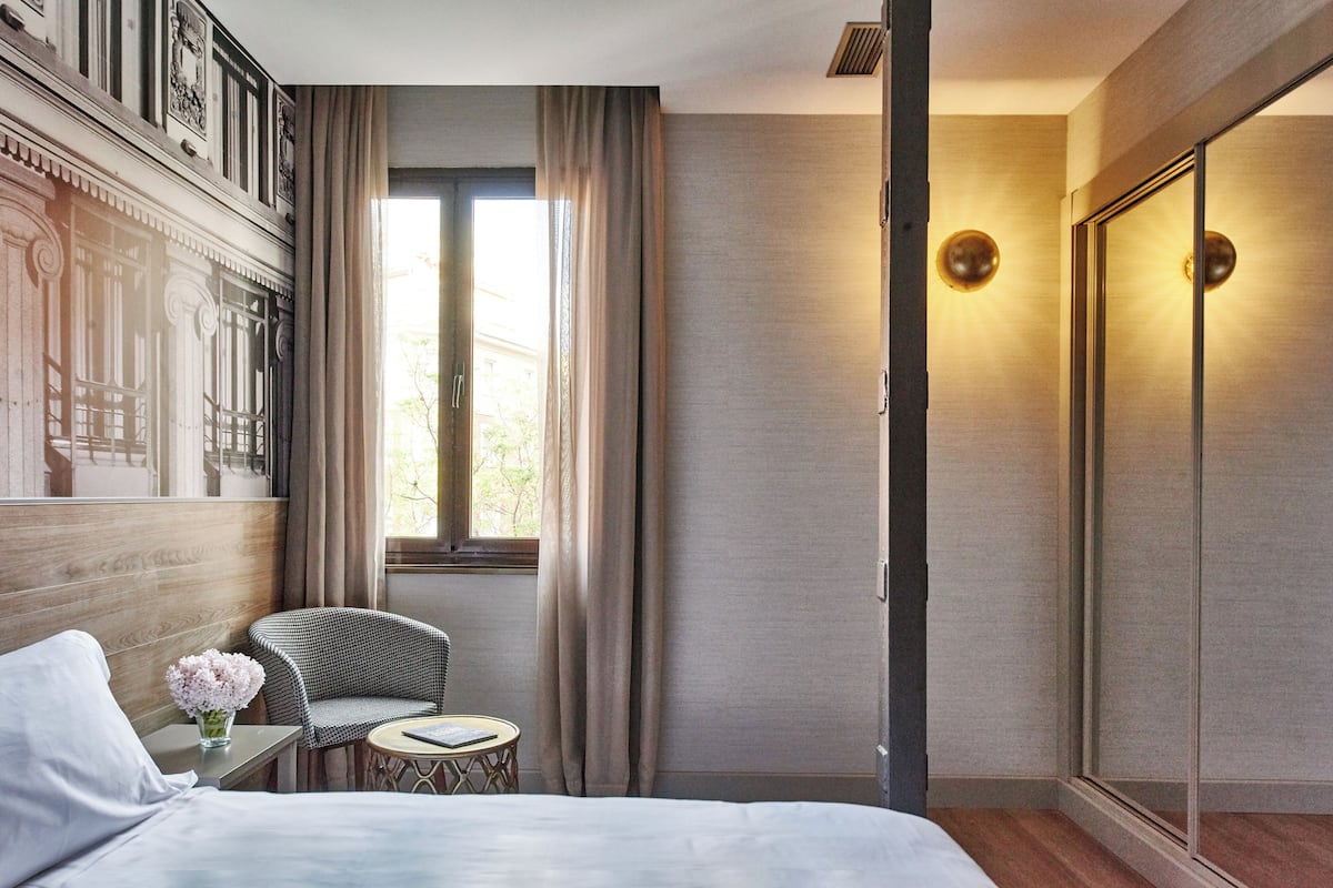 Une chambre d’hôtel avec un lit, une chaise, une petite table, un vase de fleurs, une fenêtre avec des rideaux et une armoire.