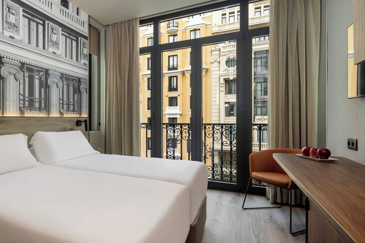 Une chambre d’hôtel avec un grand lit, un bureau et une vue sur la ville depuis un balcon.