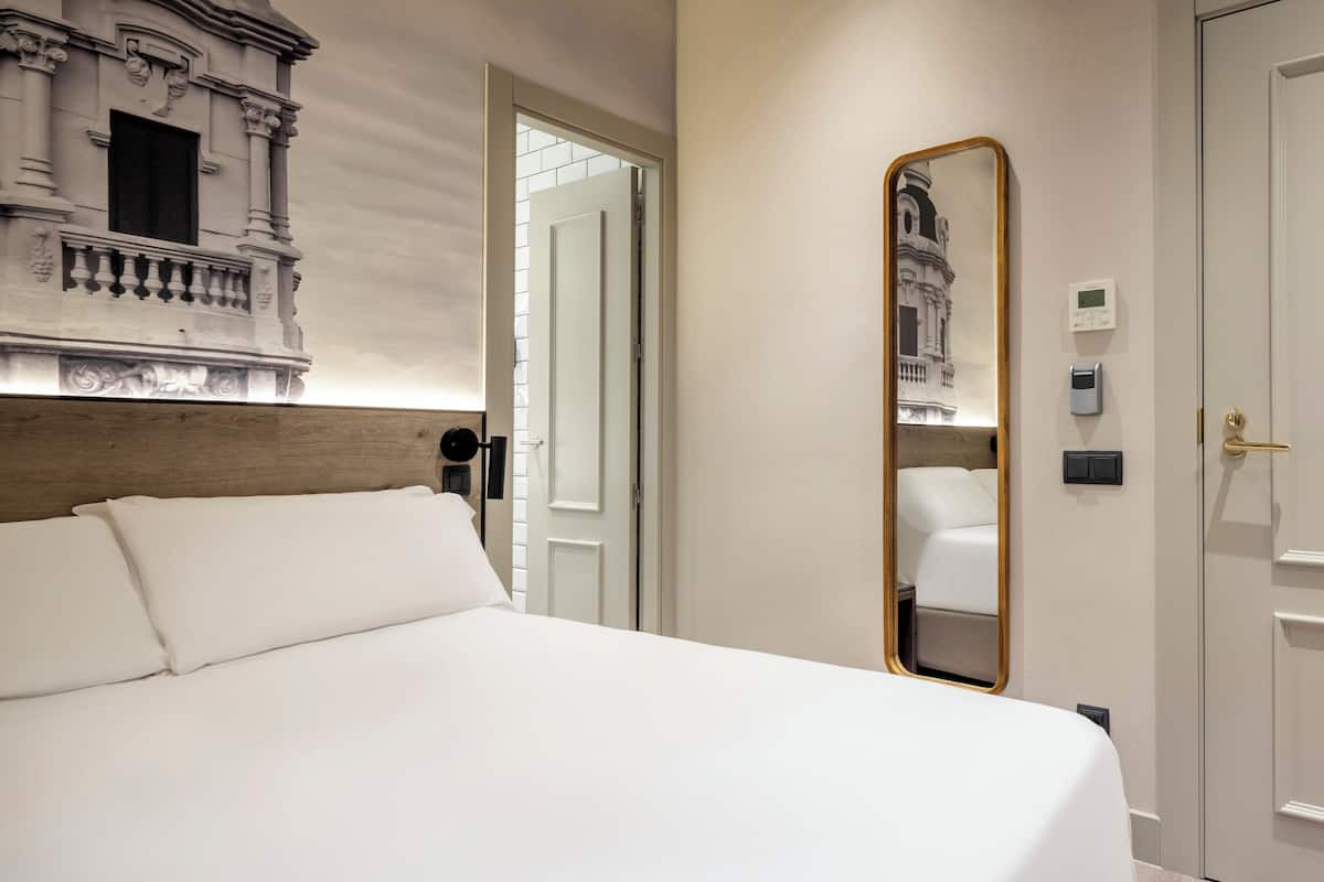 Une chambre d’hôtel avec un grand lit, une tête de lit en bois, un miroir fixé au mur et une porte donnant sur une autre pièce.