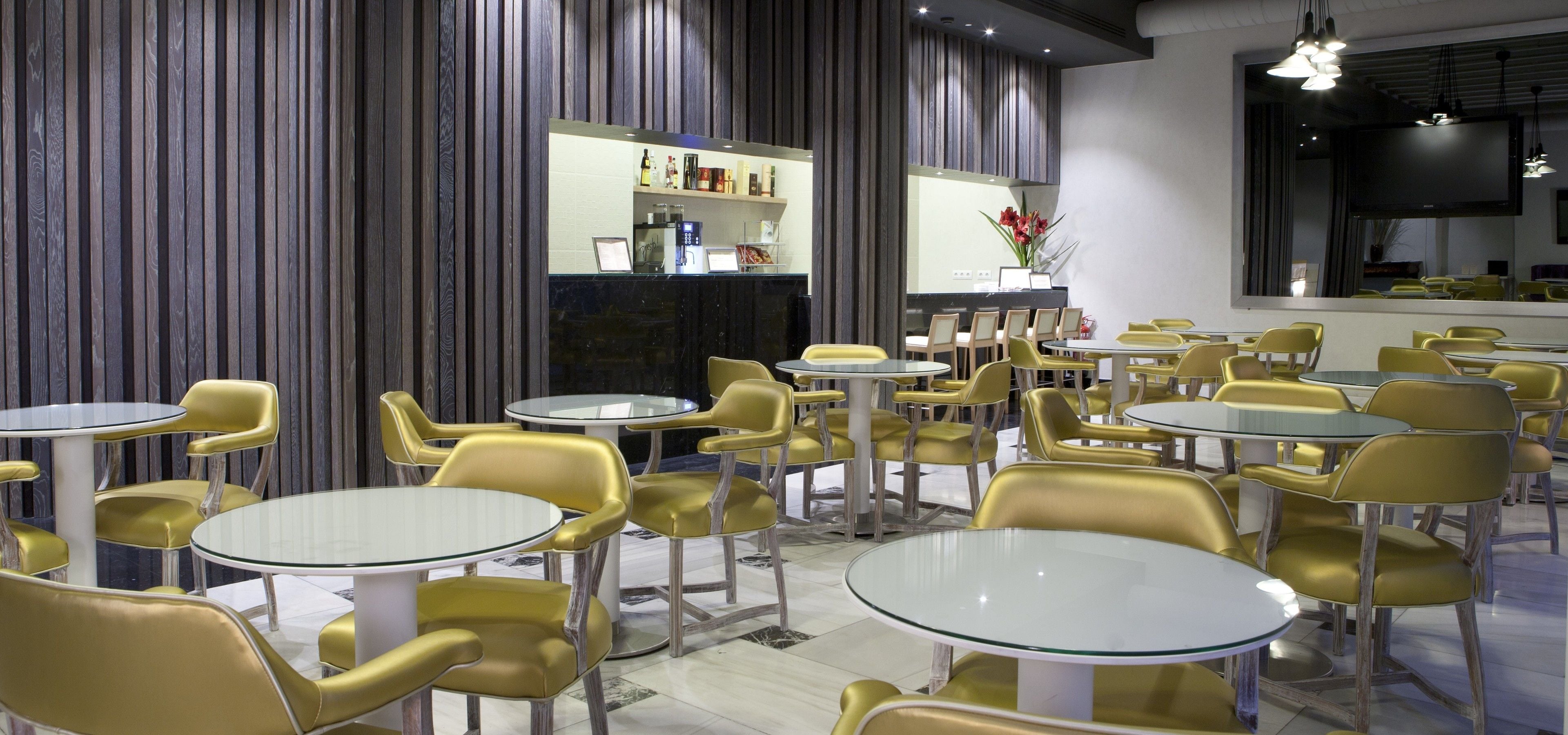 A modern restaurant interior with round tables and yellow chairs, a bar area with a counter, and a large TV screen on the wall.