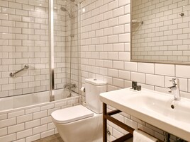 Et badeværelse med hvide fliser, en brusekabine med glasvægge, et hvidt toilet og en vask med træramme.