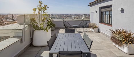 Una terraza en la azotea con mesa y sillas, plantas en macetas y vistas a la ciudad.