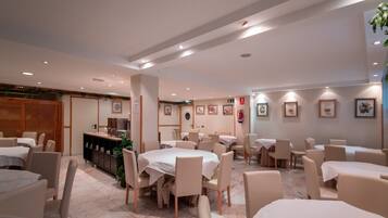 A spacious dining area with multiple tables and chairs, a kitchen area, and framed artwork on the walls.