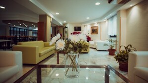 A modern lobby with a marble floor, yellow sofas, a glass coffee table with a vase of flowers, and potted plants. - Hotel Astures (Oviedo)
