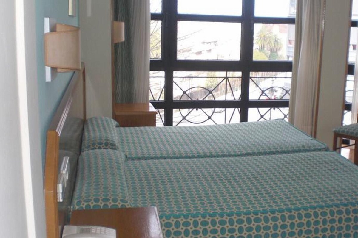 Un dormitorio con una cama, una silla, un ventanal y una lámpara.