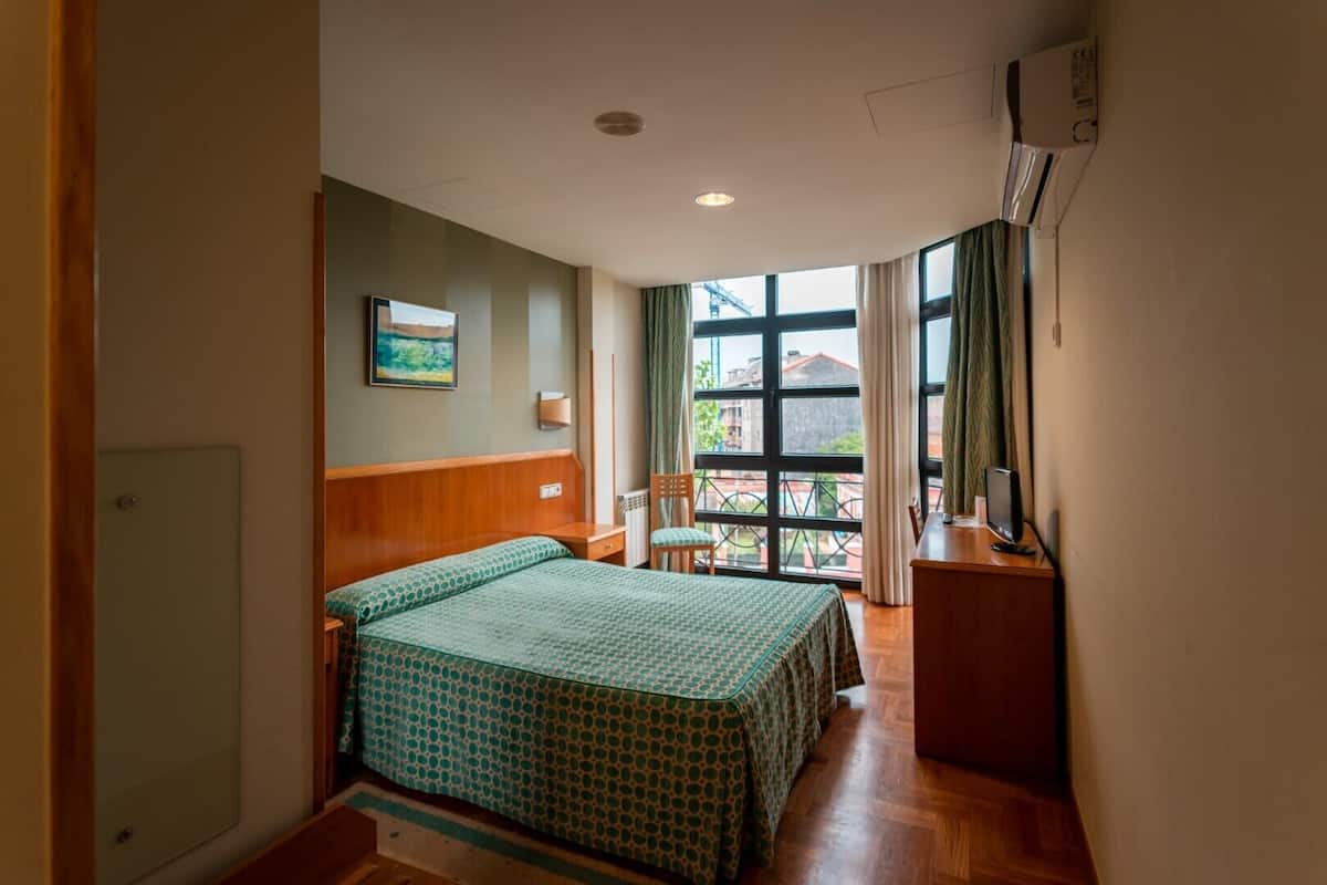 A hotel room with a bed, a desk, a chair, a window with curtains, and a painting on the wall.