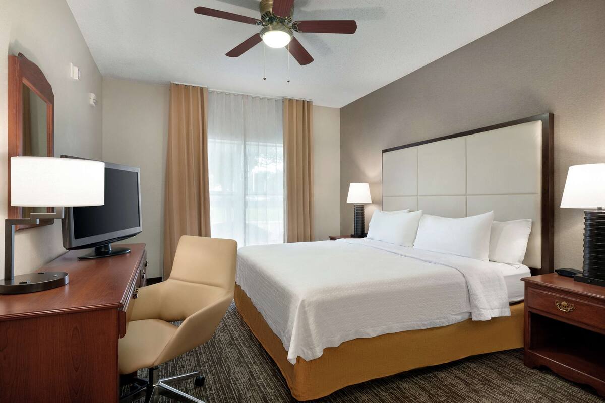 Een hotelkamer met een bed, een bureau met een televisie, een stoel, een nachtkastje met een lamp en een plafondventilator.