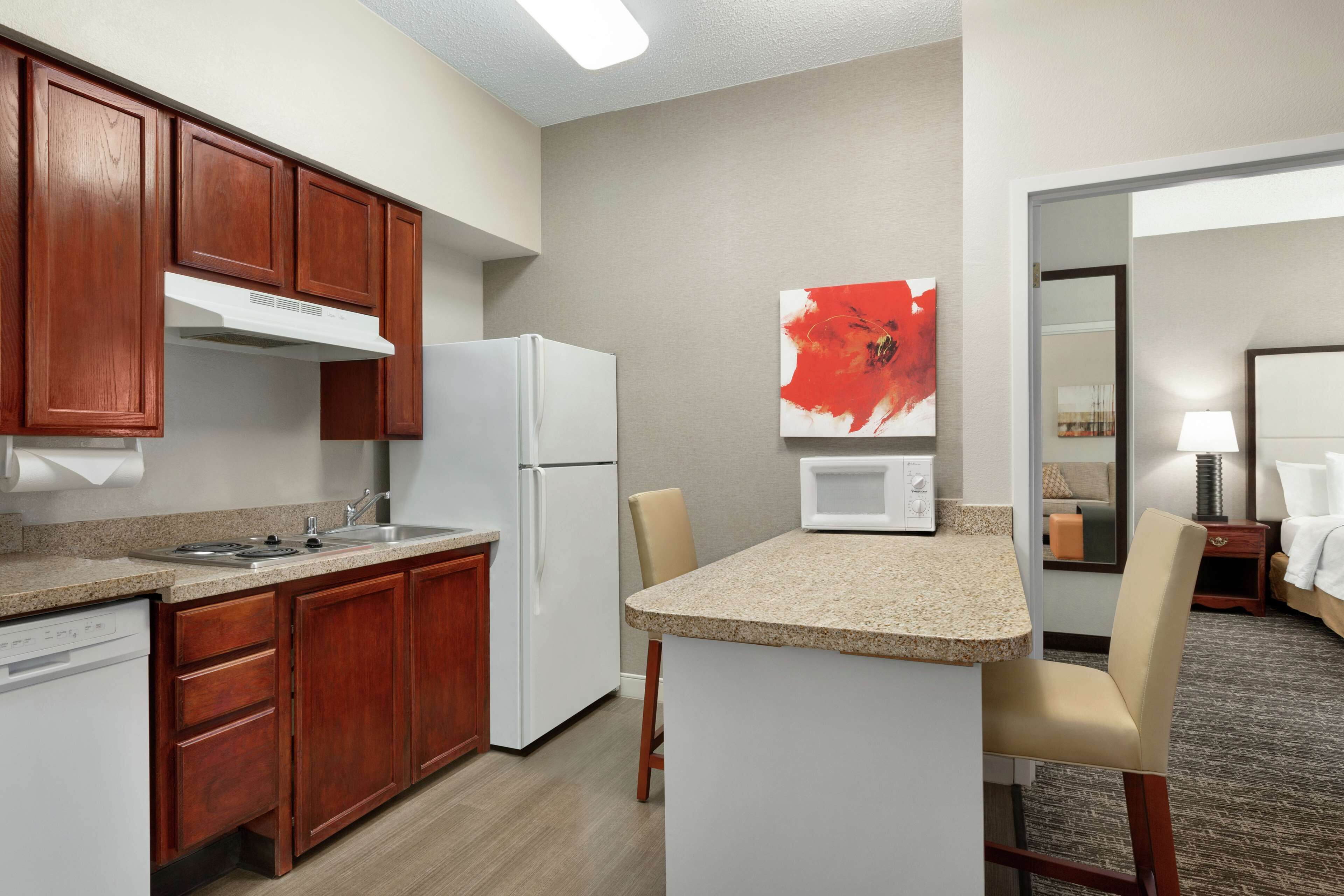 A compact hotel kitchen with wooden cabinets, a white refrigerator, a microwave, and a small island with a chair.