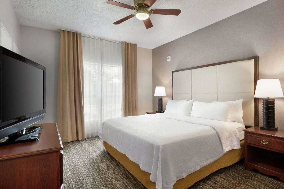 Une chambre d’hôtel avec un lit, deux lampes de chevet, un téléviseur posé sur un meuble en bois et un ventilateur de plafond.