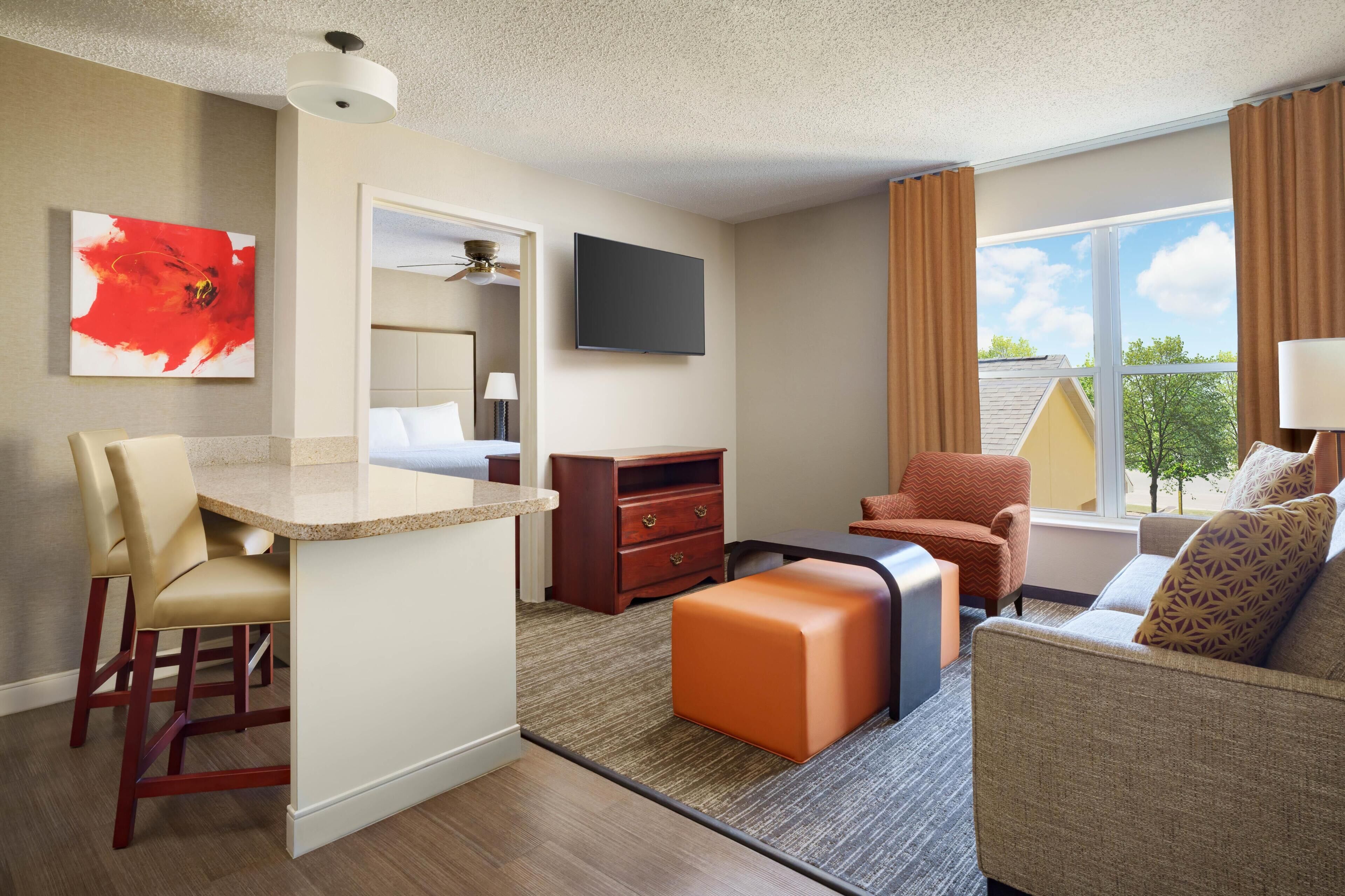 A hotel room with a kitchenette, a flat-screen TV, a sofa, a chair, and a window with a view of trees.