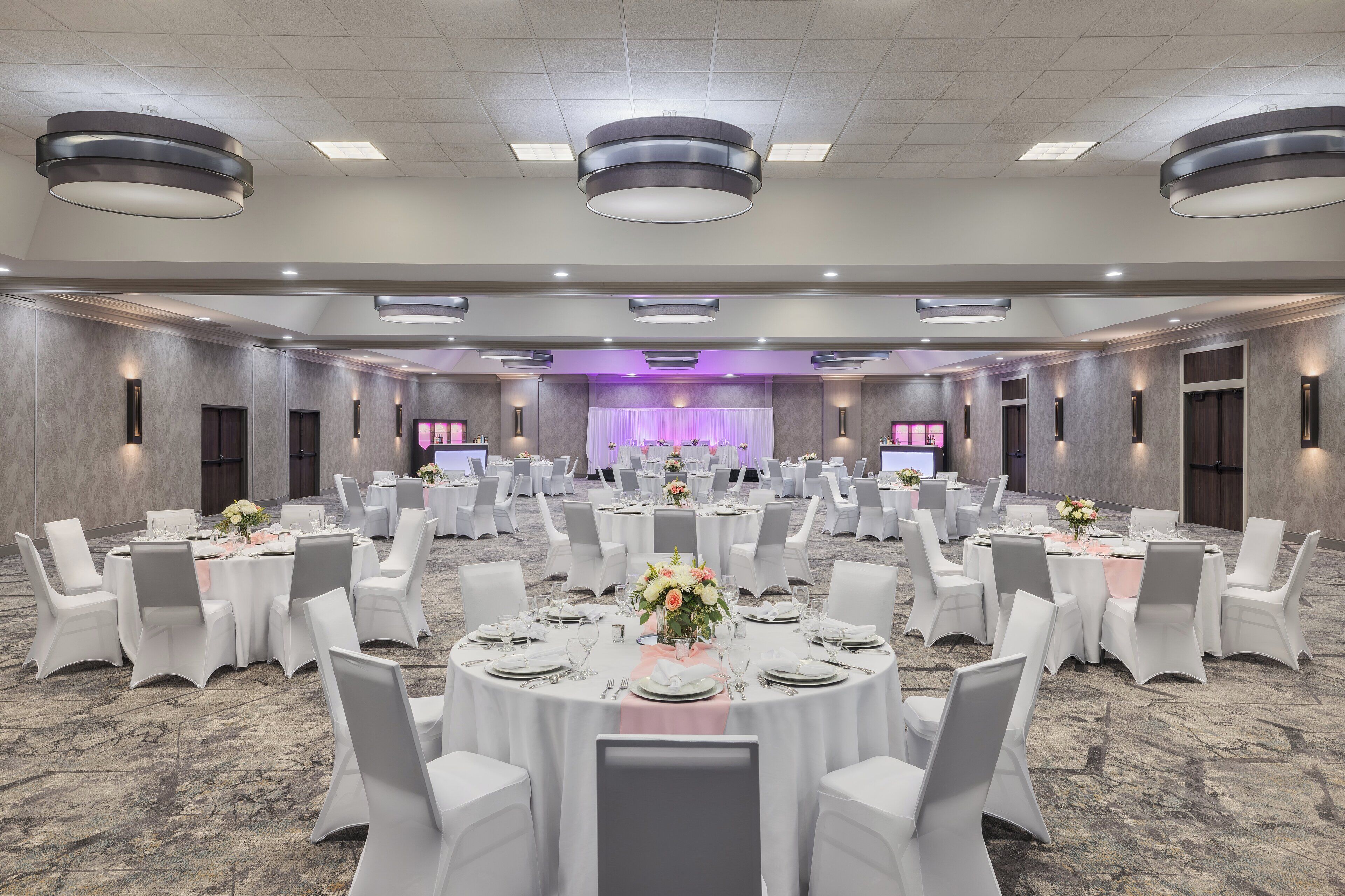 A banquet hall with round tables, white chairs, and a stage with purple lighting.