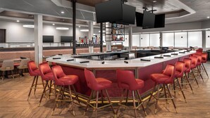 A modern bar area with a curved counter, red bar stools, and a television mounted on the ceiling. - Holiday Inn Wichita East I-35 by IHG (Wichita)