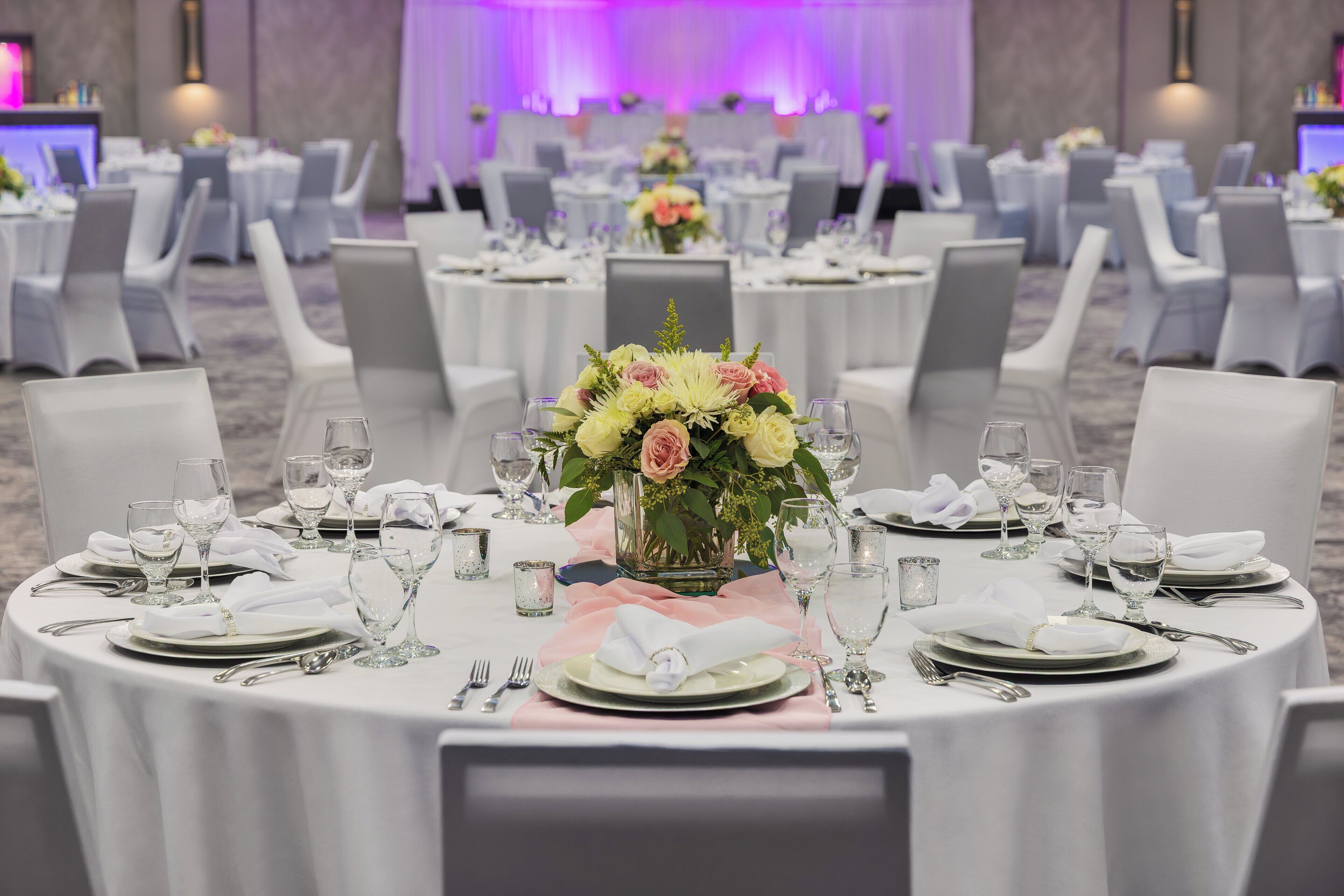 A banquet hall with round tables set for a formal event, featuring white tablecloths, neatly arranged silverware, and floral centerpieces.