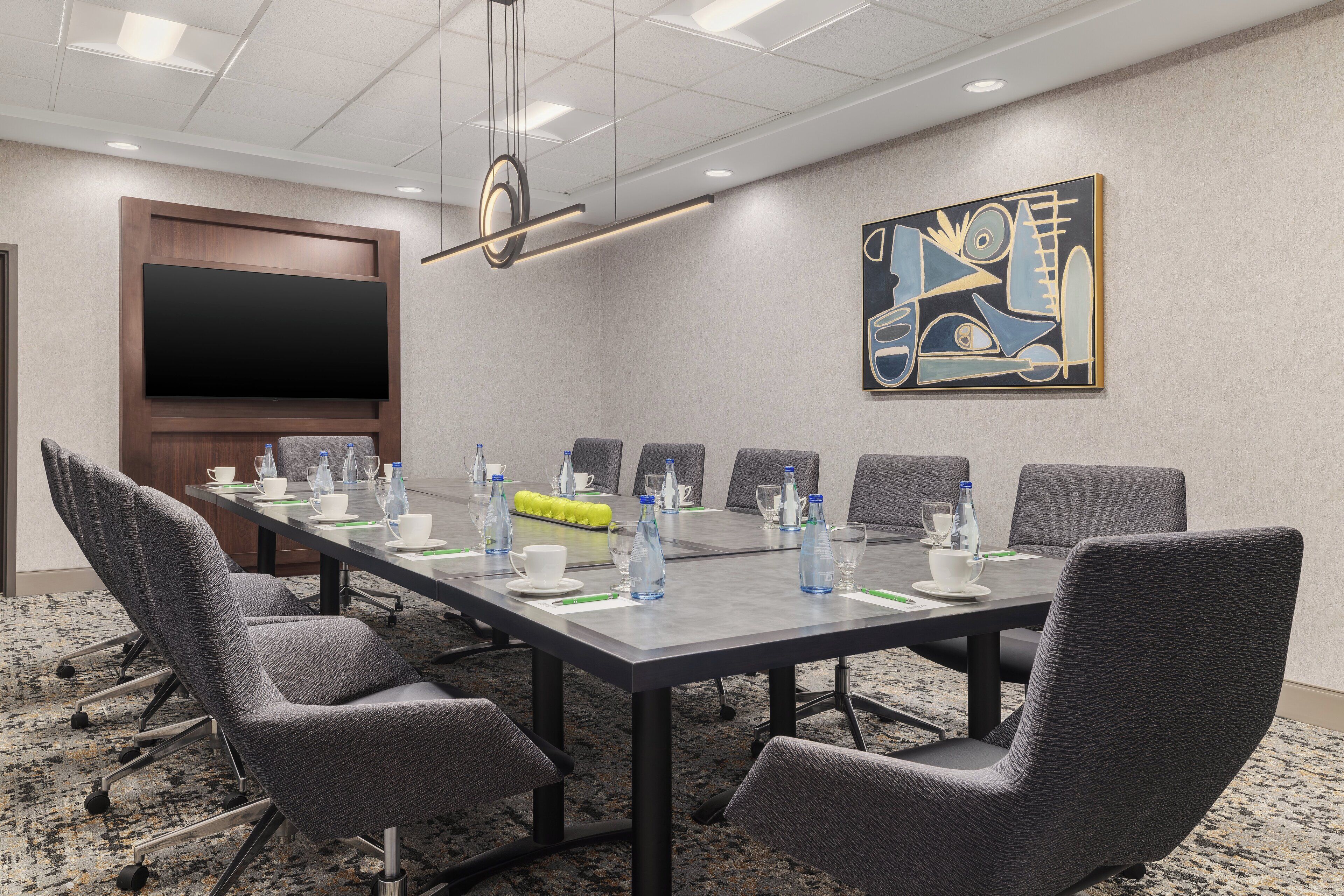 A conference room with a large table, grey chairs, a flat-screen TV, and a painting on the wall.