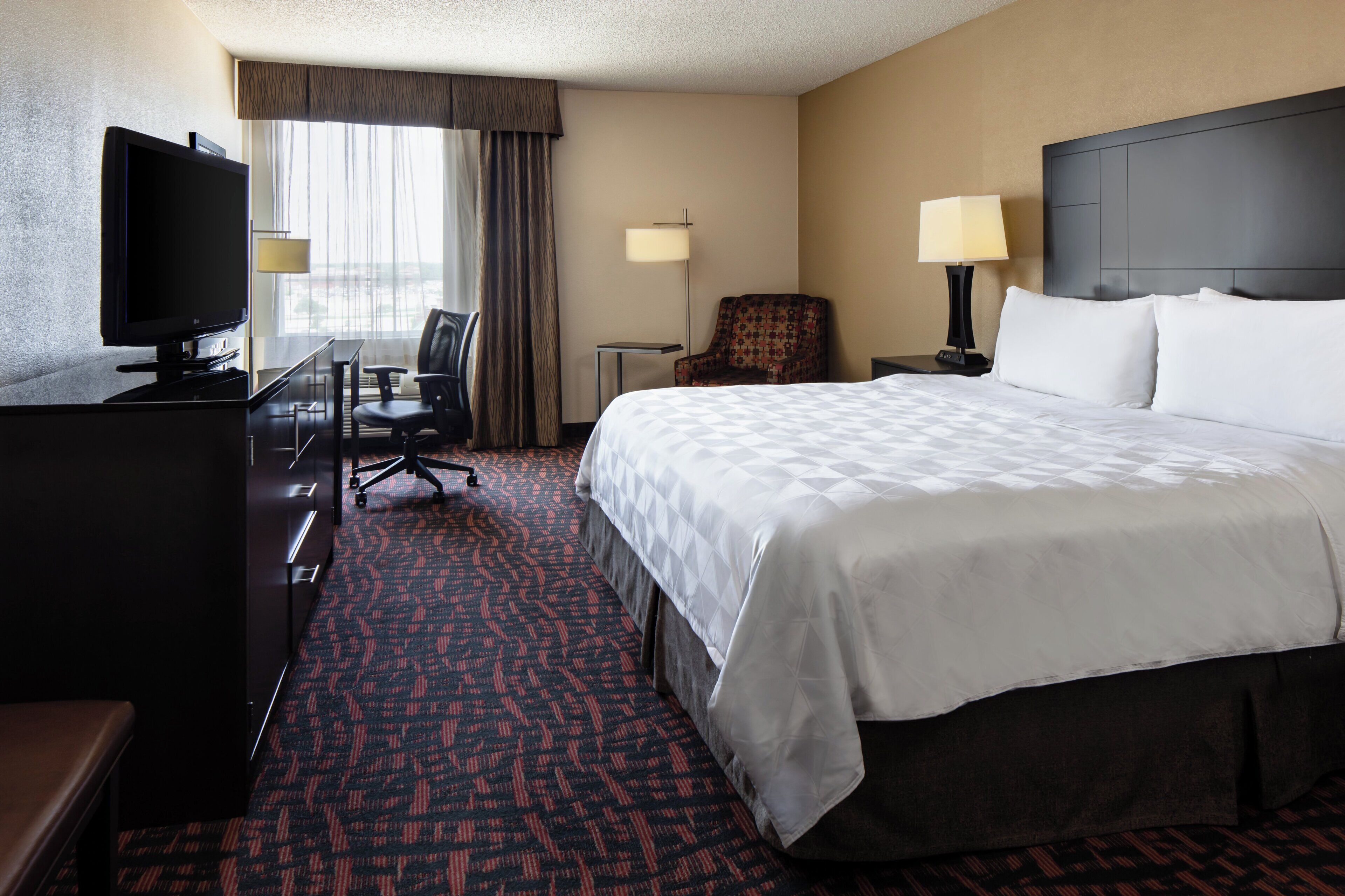 A hotel room with a bed, a desk with a TV, a chair, a lamp, and a window with curtains.